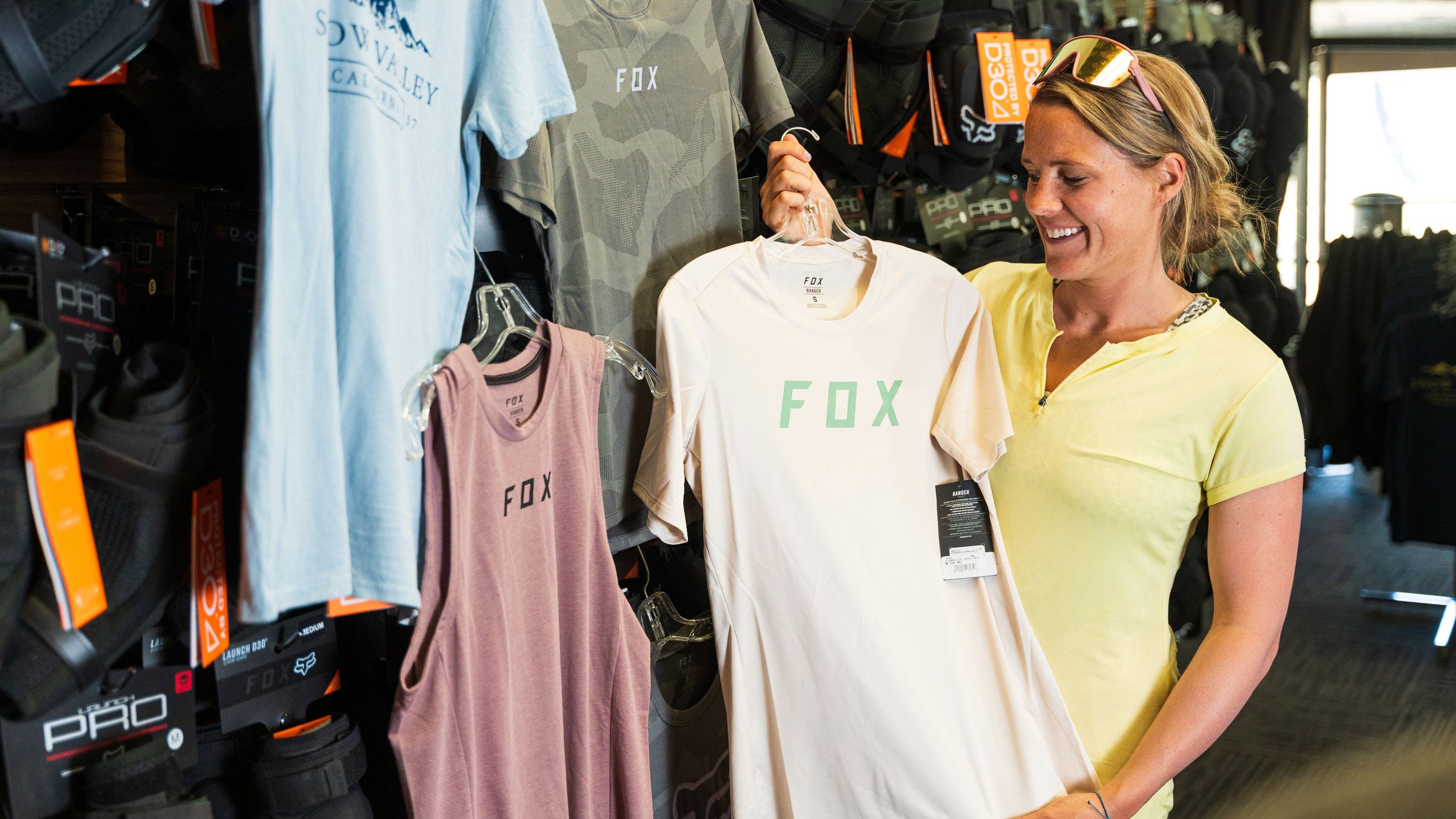 An adult holding up a beige Fox clothing shirt inside the Sports Shop at Snow Valley.