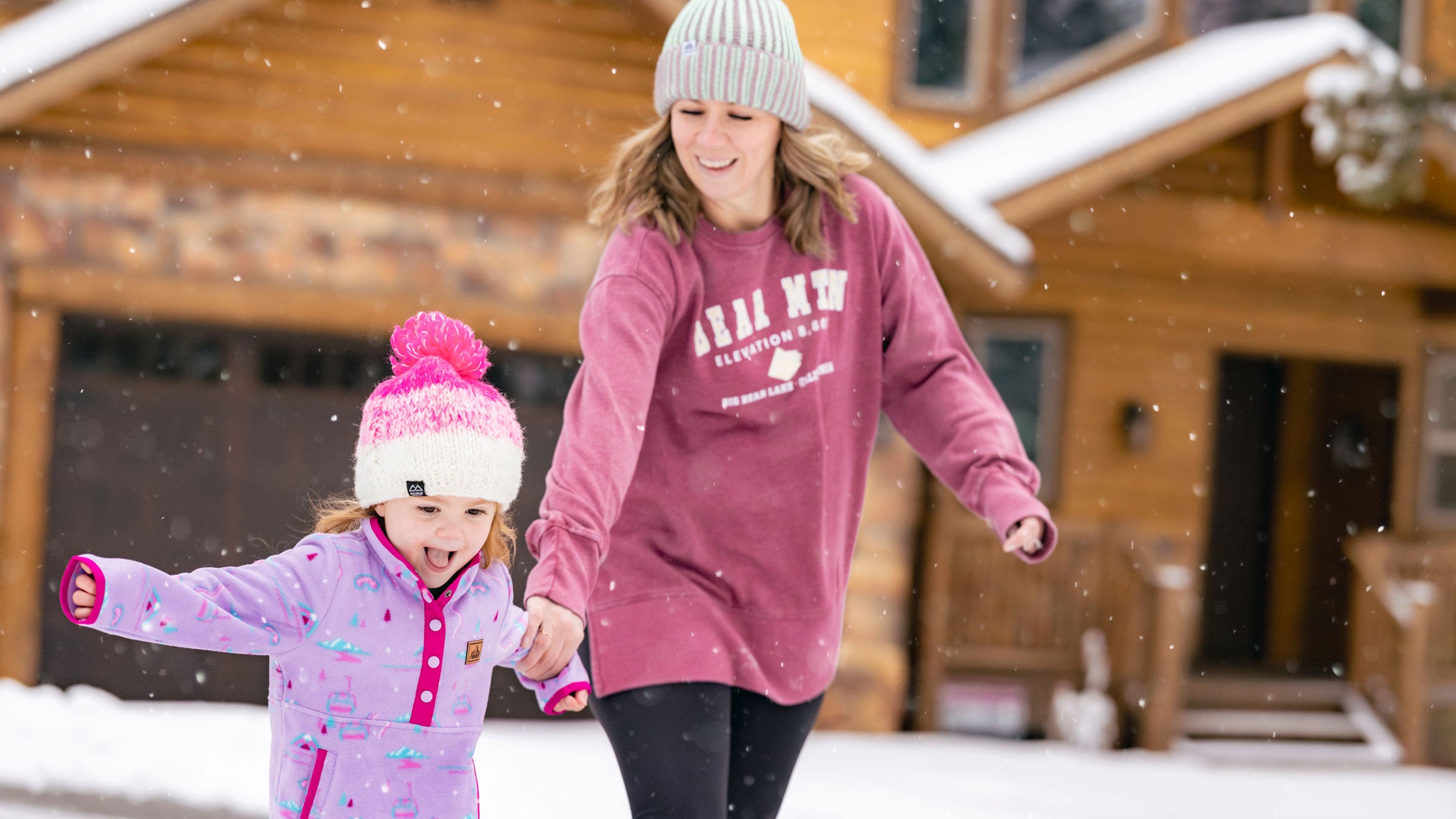An adult in a pink Bear Mountain crew neck sweatshirt and light colored beanie, holding the arm of a little kiddo wearing a purple and pink button up jacket with beanie during a snowy day in front of a cabin.