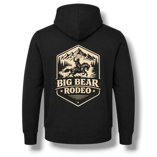 The back of a black hoodie, featuring a large white colored graphic print that reads 'BIG BEAR RODEO 2026' with an illustration of a cowboy on a horse in front of mountains.