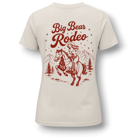 A light beige short-sleeve t-shirt with a vintage-style red graphic on the back. The graphic features the text 'Big Bear Rodeo' above an illustration of a cowgirl riding a bucking horse, with mountains and pine trees in the background.