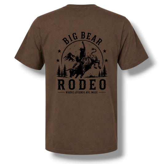 A dark color short-sleeve t-shirt with a black graphic print on the back featuring a cowboy riding a bucking bull in front of mountains and trees, with the text 'BIG BEAR RODEO' and 'WHERE LEGENDS ARE MADE'.
