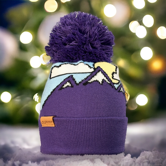 Beanie with snow capped purple mountains, clouds and a sun on a blue sky with purple pom pom and BBMR Logo tag on cuff of beanie.