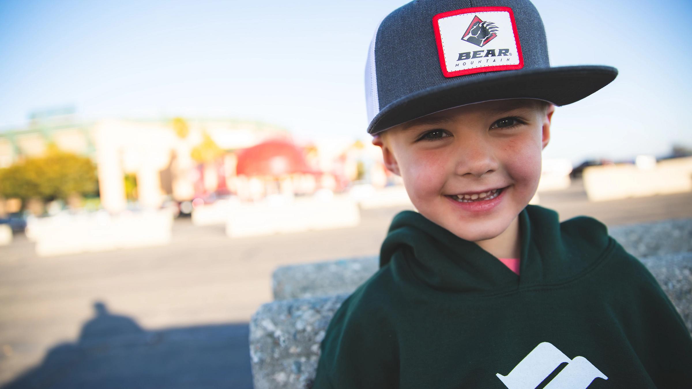 A smiling young boy wearing a dark hoodie and a dark gray/black baseball cap with a red and white 'BEAR MOUNTAIN' patch, photographed outdoors with a blurred background of Angels Stadium.