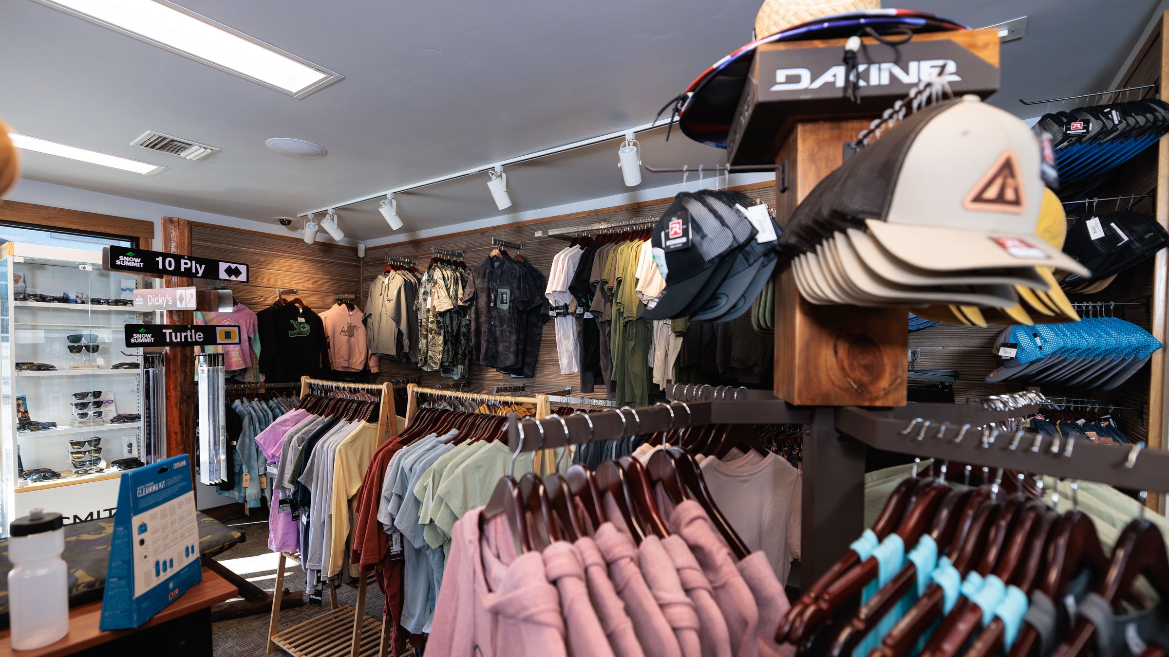 A retail store interior showing racks of clothing and a display of baseball caps, including a prominent DAKINE cap on a wooden stand.