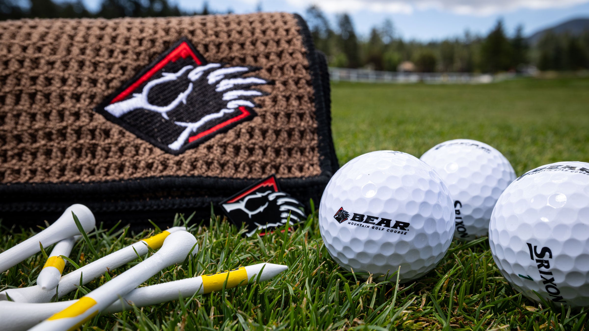 A close-up outdoor shot of Bear Mountain Golf Course golf accessories on grass, including white golf balls with visible brand logos, white tees with yellow accents, and a brown woven golf mat with a black-and-white bear paw patch.