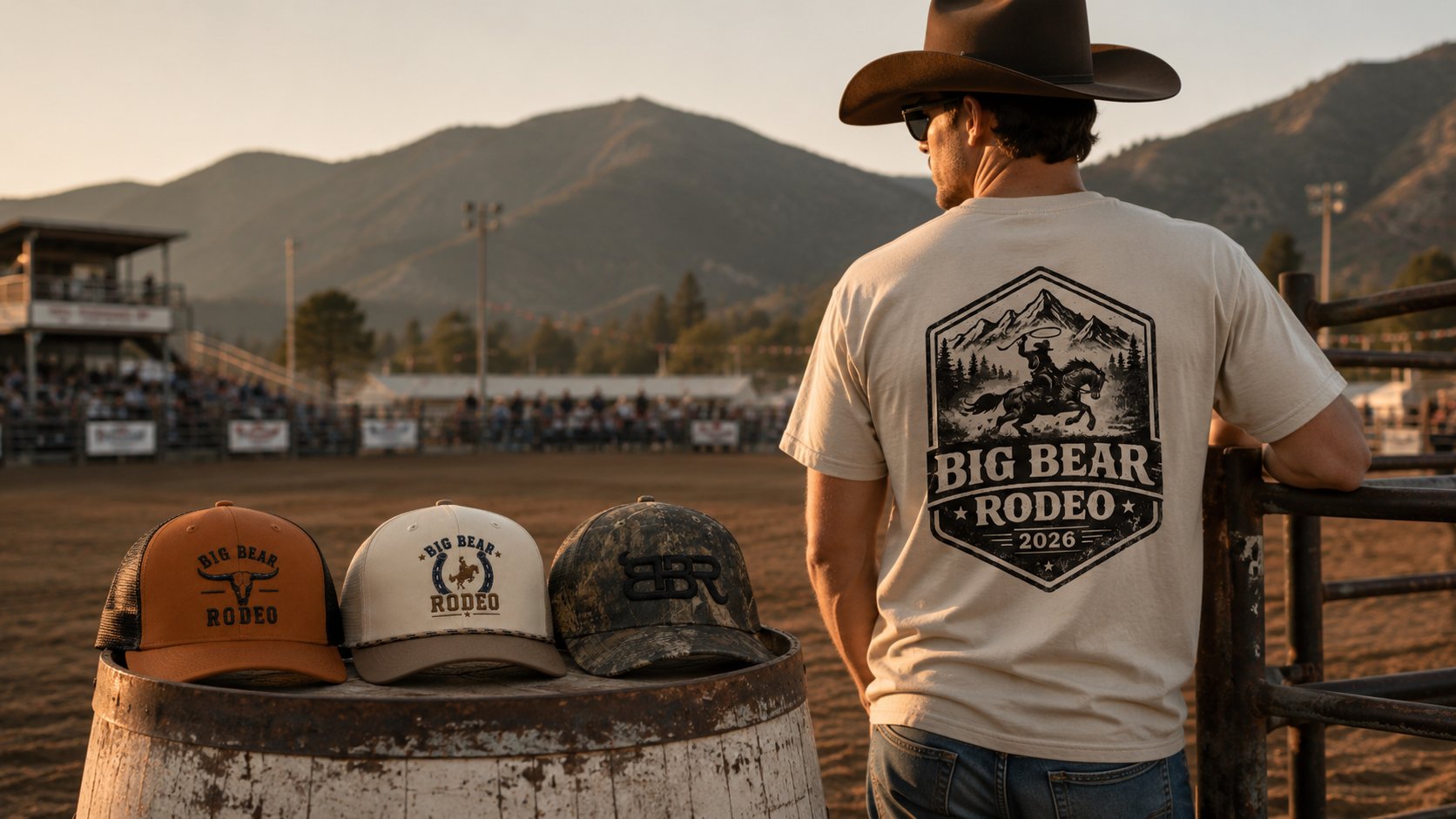 A man in a cowboy hat stands at a fence in a rodeo arena, wearing a white Big Bear Rodeo 2026 t-shirt. Three hats, with Big Bear Rodeo logos, are displayed on a barrel in the foreground against a backdrop of mountains and the arena.