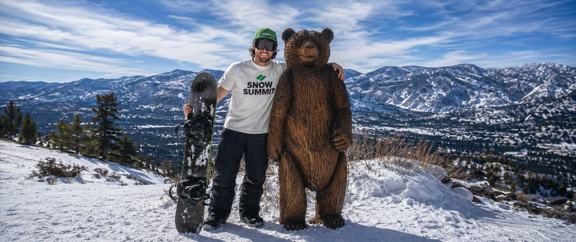 Adult, wearing a green hat with a white Snow Summit logo t-shirt. holding their snowboard at the top of the mountain posing next to a wooden bear statue on a sunny winter day.