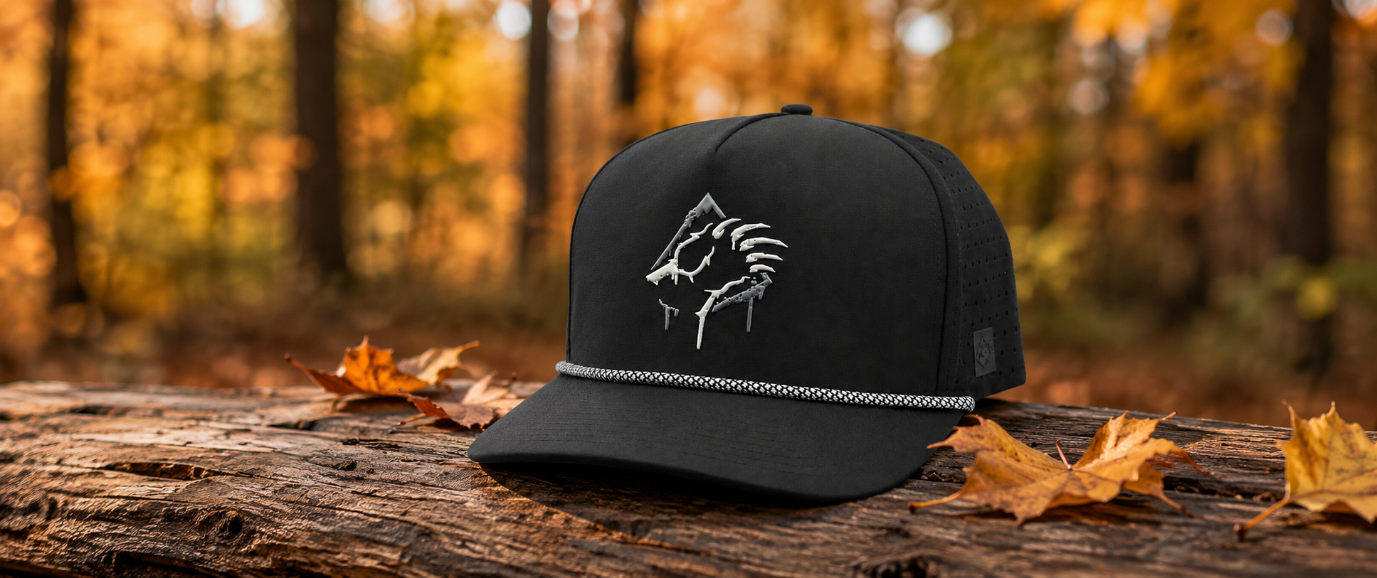 A black baseball cap with a white embroidered bear claw logo and a silver rope detail on the brim, resting on a log with autumn leaves and a blurred forest background.