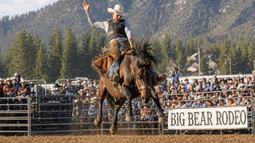 A cowboy in a white hat rides a bucking brown horse during a rodeo at Big Bear Rodeo, with a crowd and mountain backdrop.