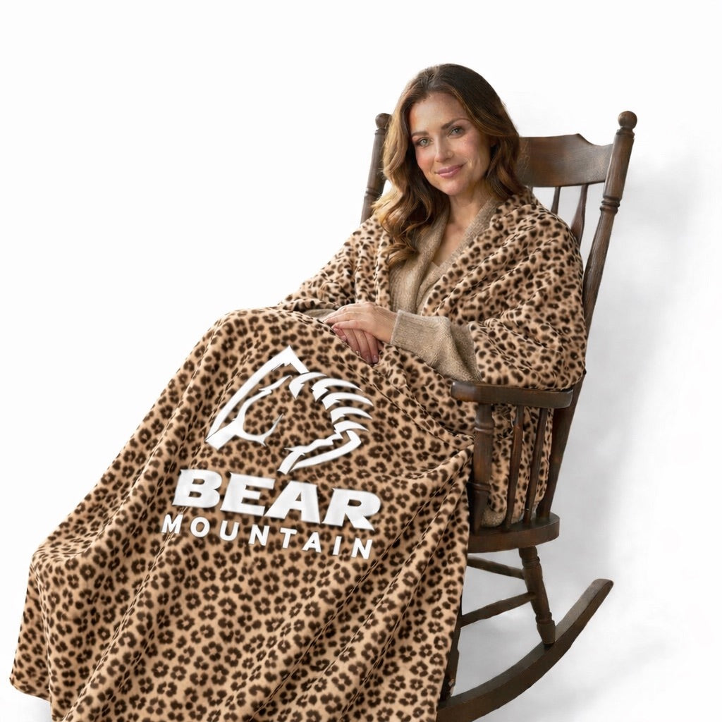 Adult with long brown hair sitting in rocking chair with a cheetah print blanket  with Bear Mountain logo draped around.
