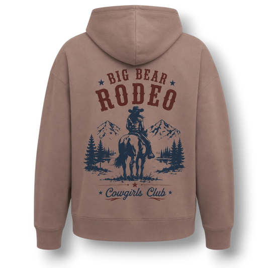 A hazy pink brownish pullover hoodie shown from the back, featuring a dark blue and maroon graphic print with the text 'BIG BEAR RODEO' and 'Cowgirls Club' surrounding an illustration of a cowgirl on a horse with mountains and pine trees.