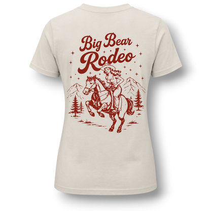A light beige short-sleeve t-shirt with a vintage-style red graphic on the back. The graphic features the text 'Big Bear Rodeo' above an illustration of a cowgirl riding a bucking horse, with mountains and pine trees in the background.