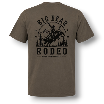 A dark color short-sleeve t-shirt with a black graphic print on the back featuring a cowboy riding a bucking bull in front of mountains and trees, with the text 'BIG BEAR RODEO' and 'WHERE LEGENDS ARE MADE'.