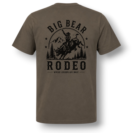 A dark color short-sleeve t-shirt with a black graphic print on the back featuring a cowboy riding a bucking bull in front of mountains and trees, with the text 'BIG BEAR RODEO' and 'WHERE LEGENDS ARE MADE'.