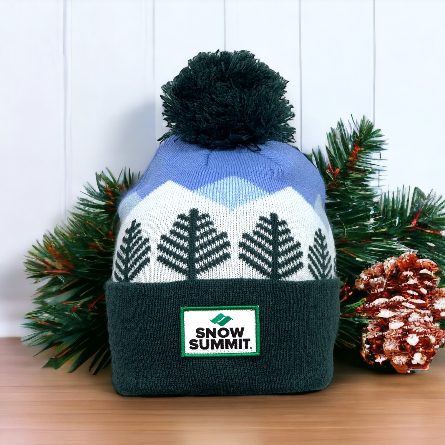 Beanie portraying stitching of trees and a blue stitched sky with snow summit logo patch on cuff of beanie and dark green pom pom on top.