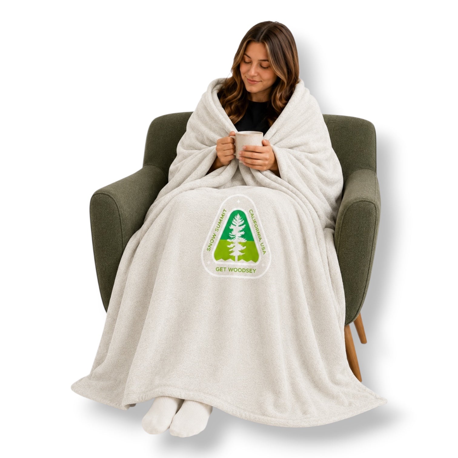 Woman sitting in a chair wrapped in a light grey blanket with a Snow Summit get woodsy logo, holding a mug.