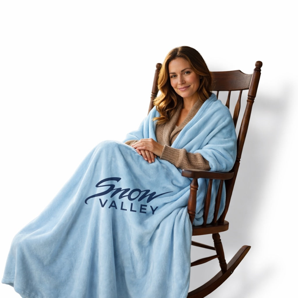 Adult with long dark hair sitting on a rocking chair wearing a blue blanket with Snow Valley branding.